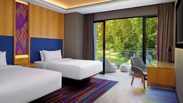 Een moderne hotelkamer met twee bedden, een bureau, een stoel en een balkon met uitzicht op groen.
