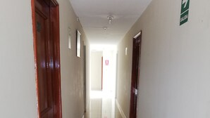 A long, narrow hallway with white tiled floors and beige walls. There are doors on both sides, and a green EXIT sign is visible on the right wall. - La Quinta de Allison Hotel & Backpackers (Lima)