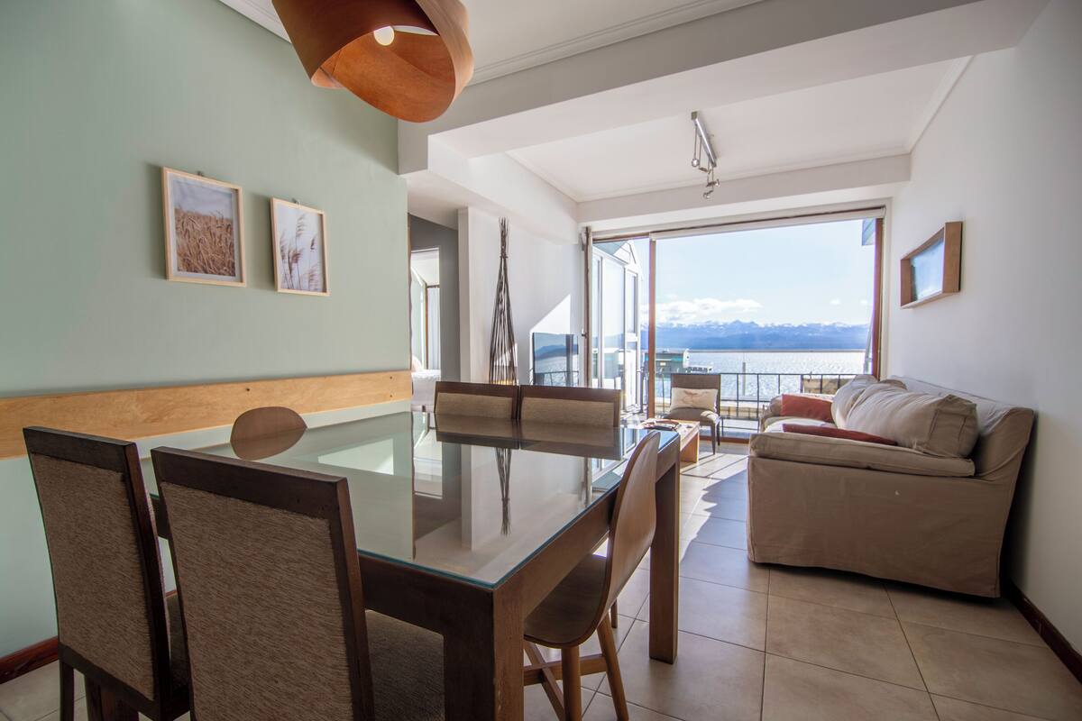 Uma área de refeições com mesa e cadeiras de vidro, um sofá e vista para montanhas através de grandes janelas.