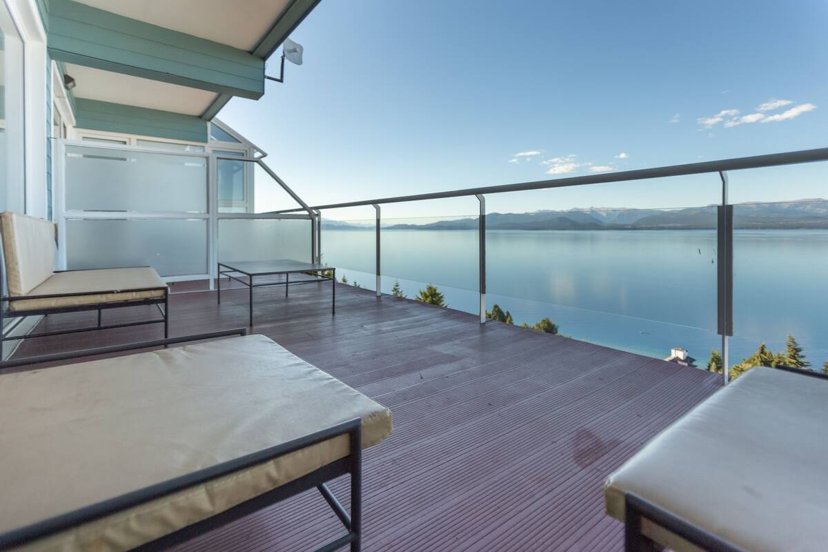 Varanda com duas cadeiras e uma mesa, com vista para um lago tranquilo e montanhas ao longe.