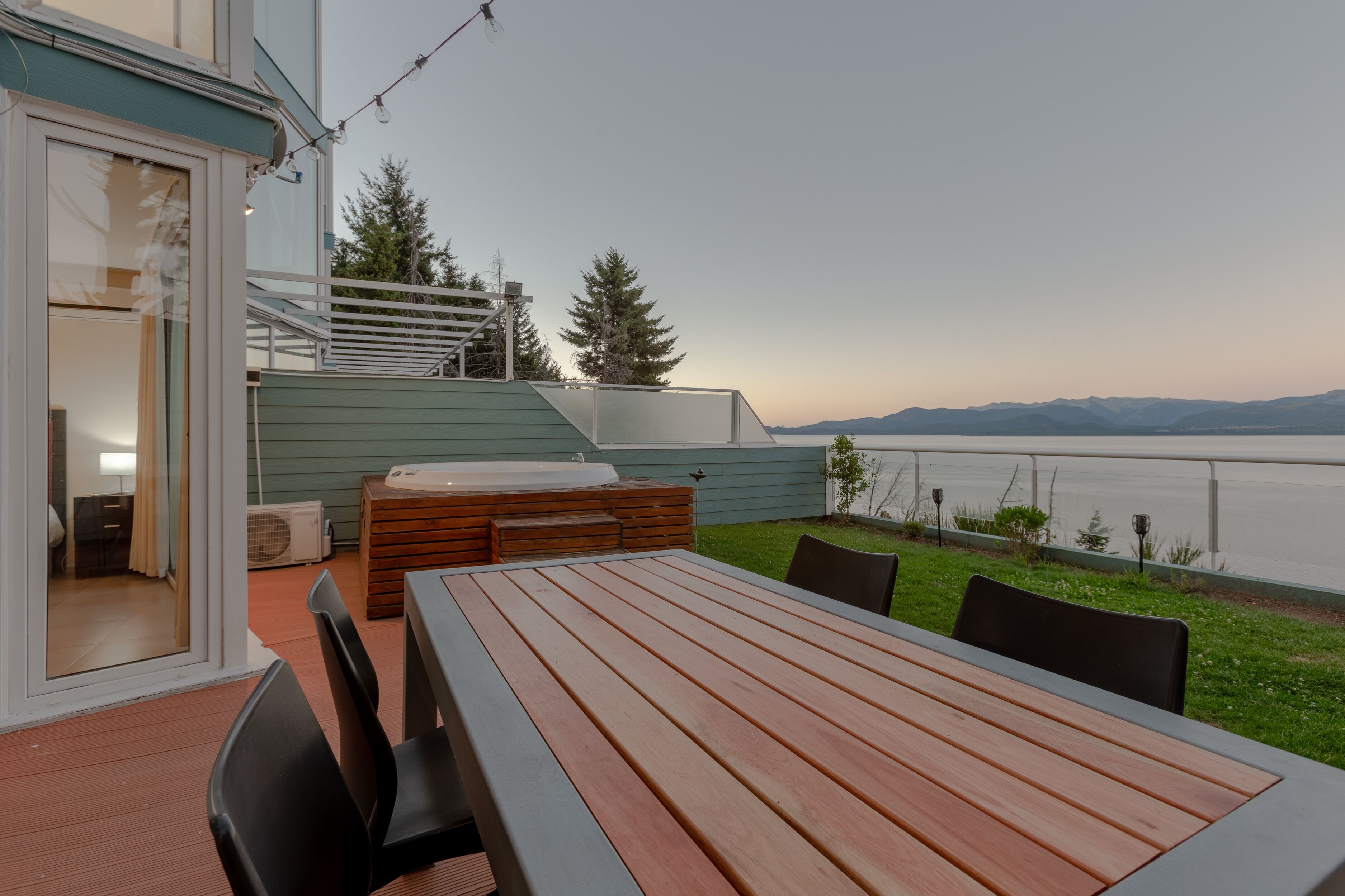 A wooden deck with a table and chairs, a hot tub, and a view of a lake and mountains.