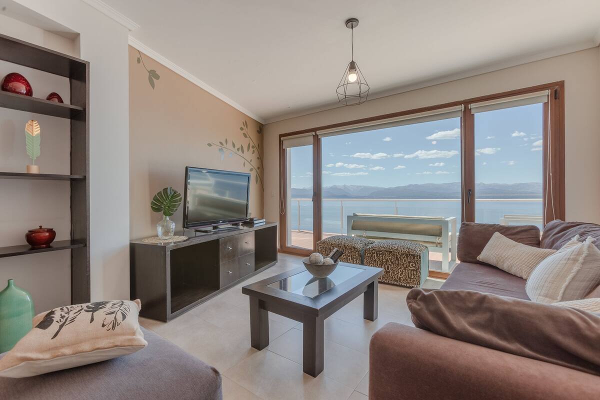Uma sala de estar moderna com televisão de tela plana, uma mesa de centro de vidro, um sofá e vista para o mar através de grandes janelas.