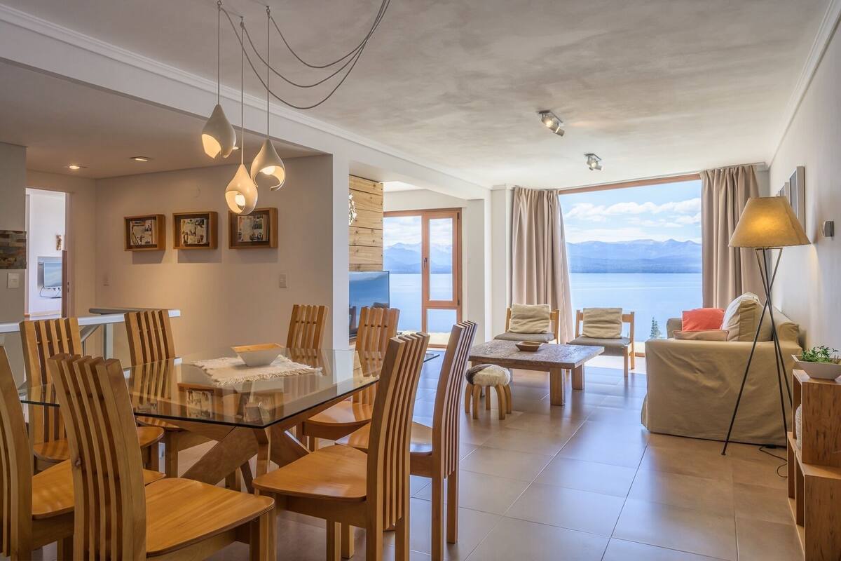 Uma sala de estar espaçosa com uma mesa de jantar de vidro, cadeiras de madeira, um sofá e vista para um lago.