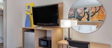 A hotel room with a flat-screen TV, a microwave, a mini fridge, a desk with a chair, a lamp, and artwork on the wall.
