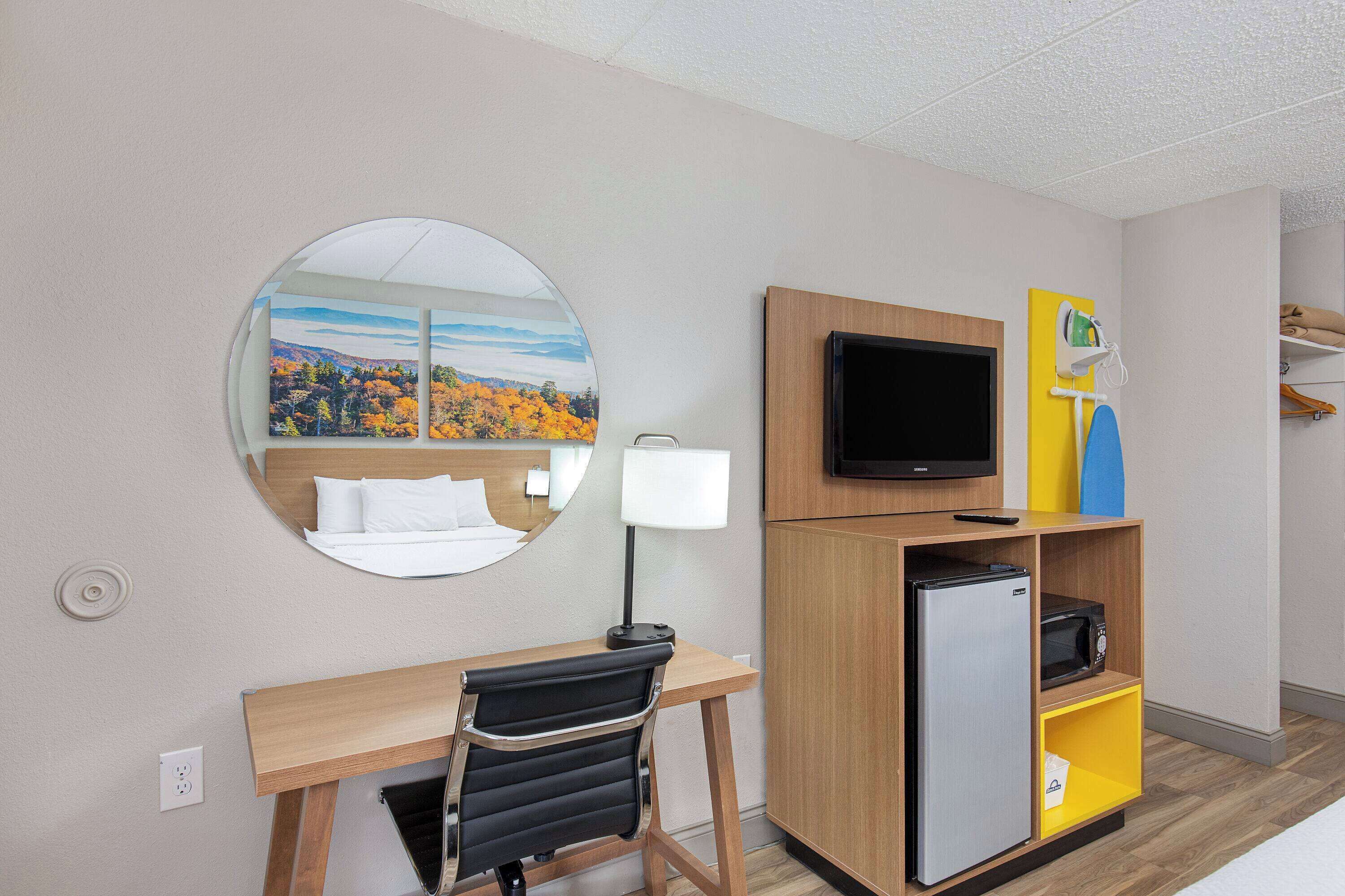 Habitación de hotel con un escritorio de madera, una silla negra, un espejo, un televisor de pantalla plana, un refrigerador y un microondas.