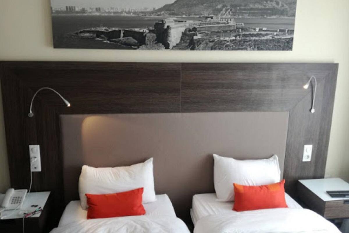 A hotel room with two beds, each with white bedding and red pillows, a large framed picture above the beds, and a dark wood headboard.