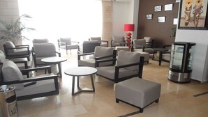 Rebedor d'hotel amb diverses zones d'estar, incloses sofàs i cadires, disposades al voltant de taules petites. Hi ha una màquina de cafè i una làmpada decorativa al centre.