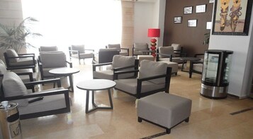 Un vestíbulo de hotel con varias zonas de estar, que incluyen sofás y sillones, distribuidos alrededor de mesitas. Hay una máquina de café y una lámpara decorativa en el centro.