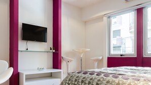 Ein Schlafzimmer mit einem Bett, einem an der Wand montierten Fernseher, einem weißen Schreibtisch, einem Sessel und einem Fenster mit Blick auf Gebäude.