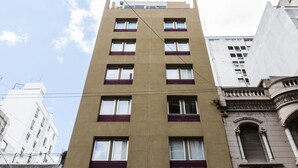 Ein mehrstöckiges Gebäude mit einer beige farbenen Fassade, zahlreichen Fenstern mit roten Rollläden und einem klassischen architektonischen Detail auf der rechten Seite.