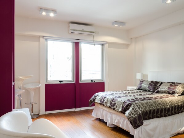 A bedroom with a bed, a window, a bedside table, a chair, and an air conditioning unit.