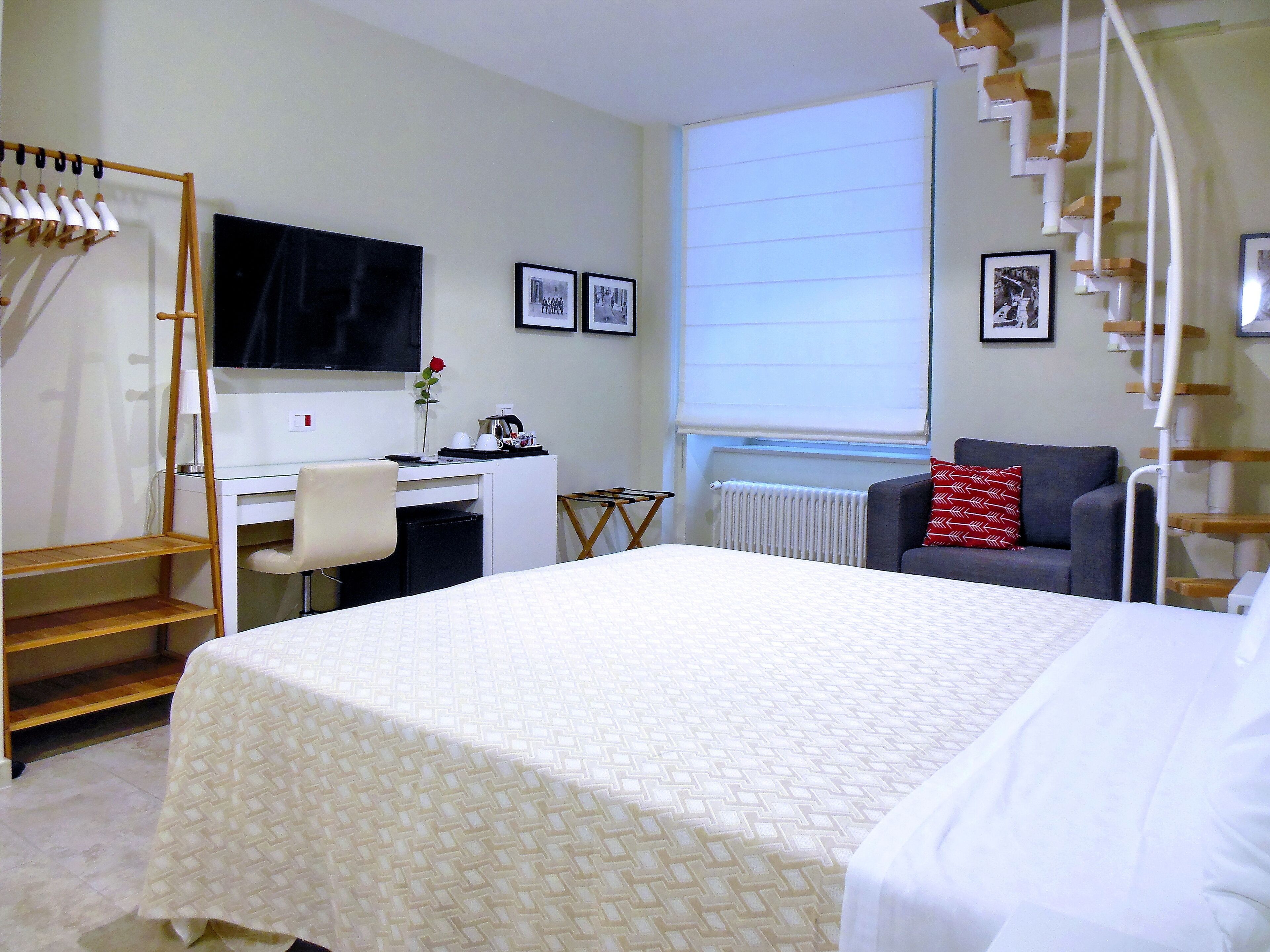 A hotel room with a bed, a desk, a chair, a radiator, a window with blinds, and a TV on the wall.