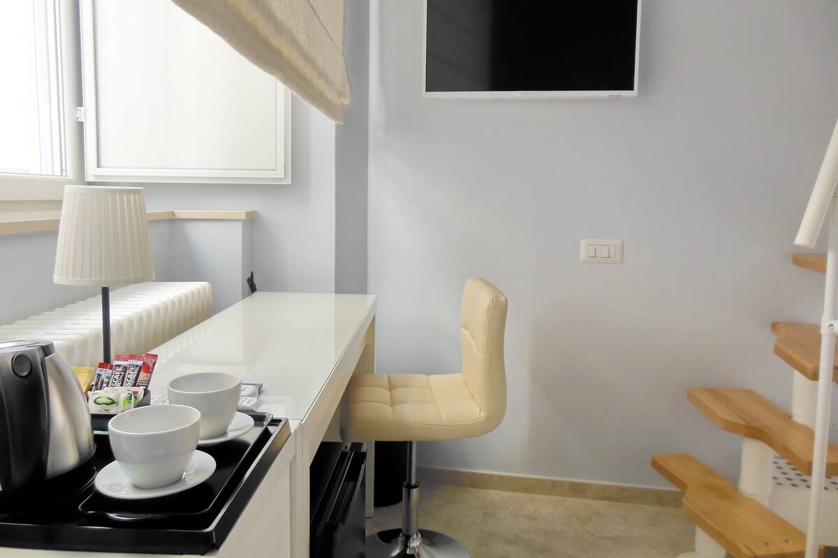 A small kitchenette with a stove, kettle, and cups on a tray. There is a white chair and a TV mounted on the wall.