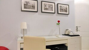 Una habitación con un escritorio, una silla, una lámpara, un jarrón con una rosa y tres cuadros en la pared.