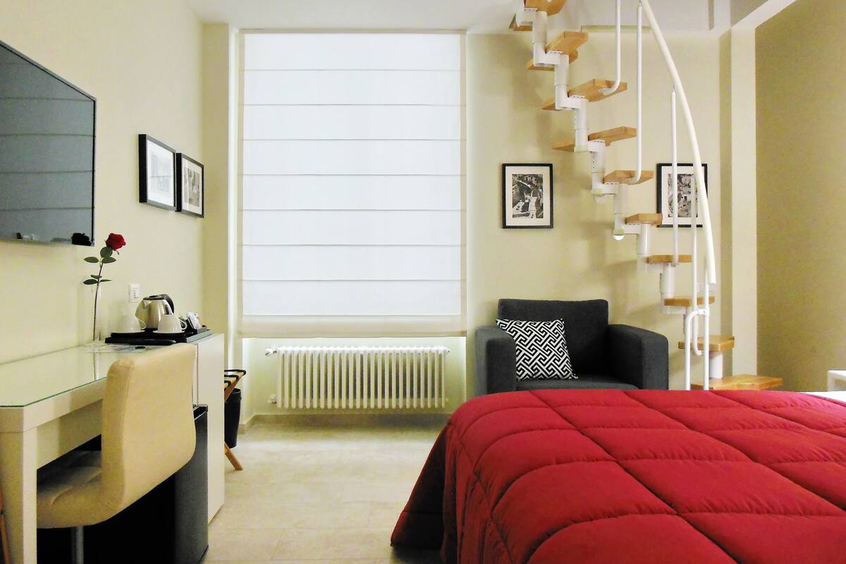 A hotel room with a bed, a desk, a chair, a radiator, a window with blinds, and a staircase with a railing.