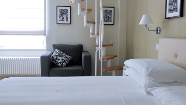 Una habitación de hotel con una cama, una silla con un cojín estampado, un radiador blanco y dos cuadros en la pared.