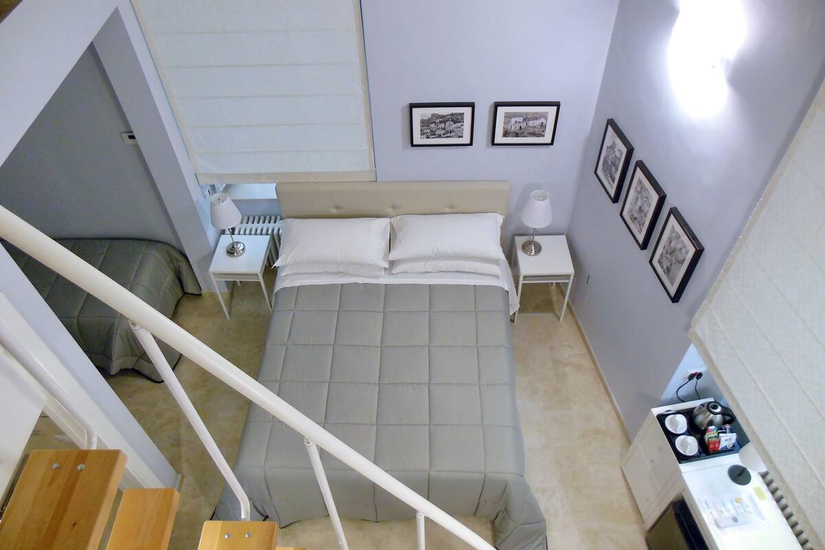 A bedroom with a bed, two bedside tables, a nightstand, a lamp, and a small kitchenette.