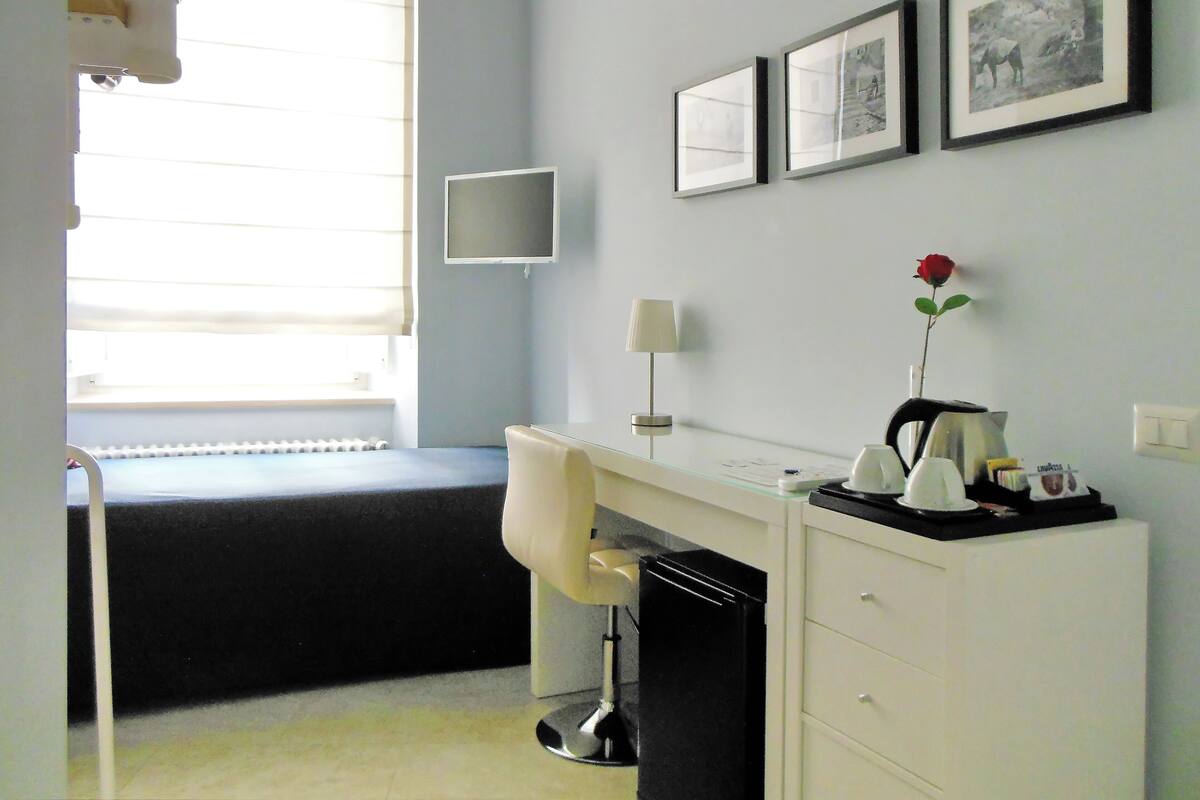 A bedroom with a bed, a desk with a chair, a lamp, a picture frame, and a rose on the desk.