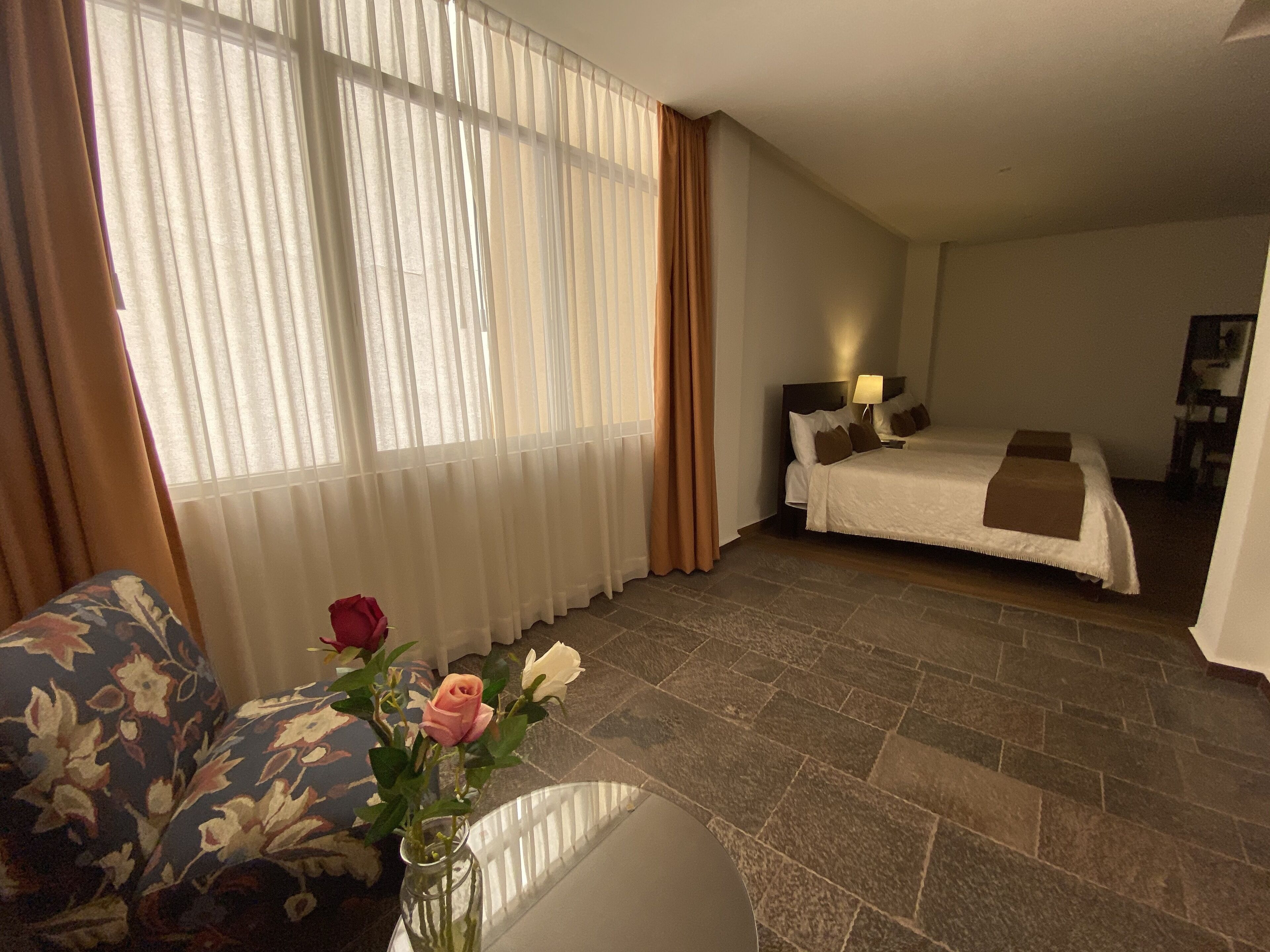 Ett hotellrum med en säng, ett nattduksbord, en lampa, en vas med blommor och ett fönster med gardiner.