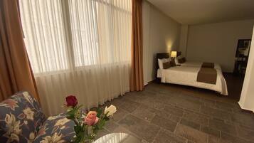 Ett hotellrum med en säng, ett nattduksbord, en lampa, en vas med blommor och ett fönster med gardiner.
