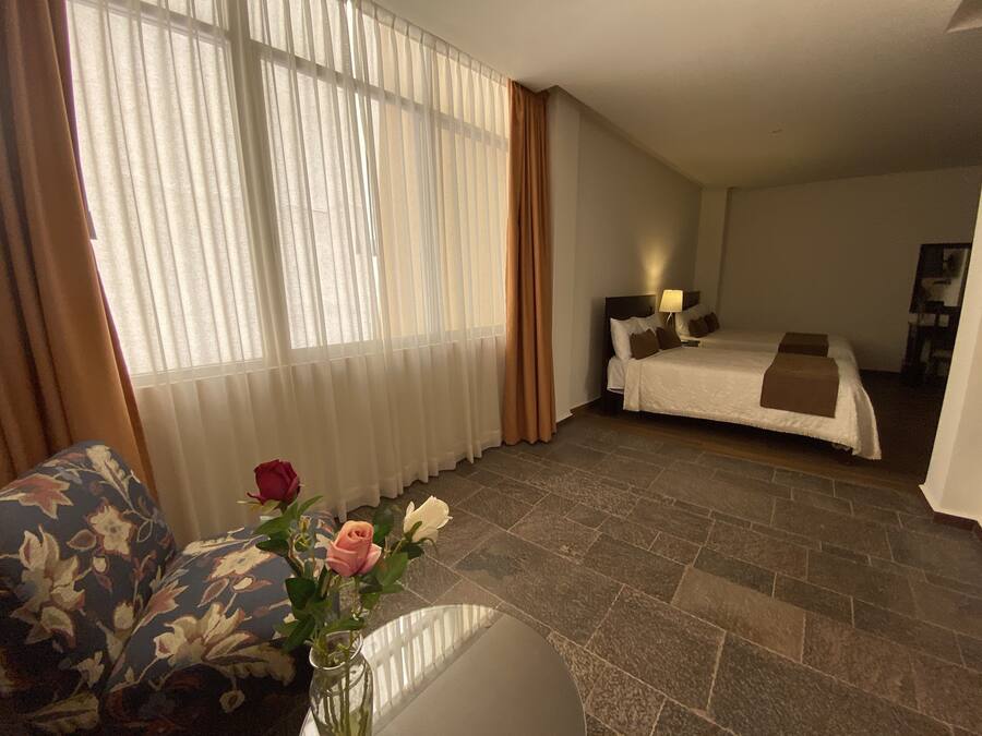 Ett hotellrum med en säng, ett nattduksbord, en lampa, en vas med blommor och ett fönster med gardiner.