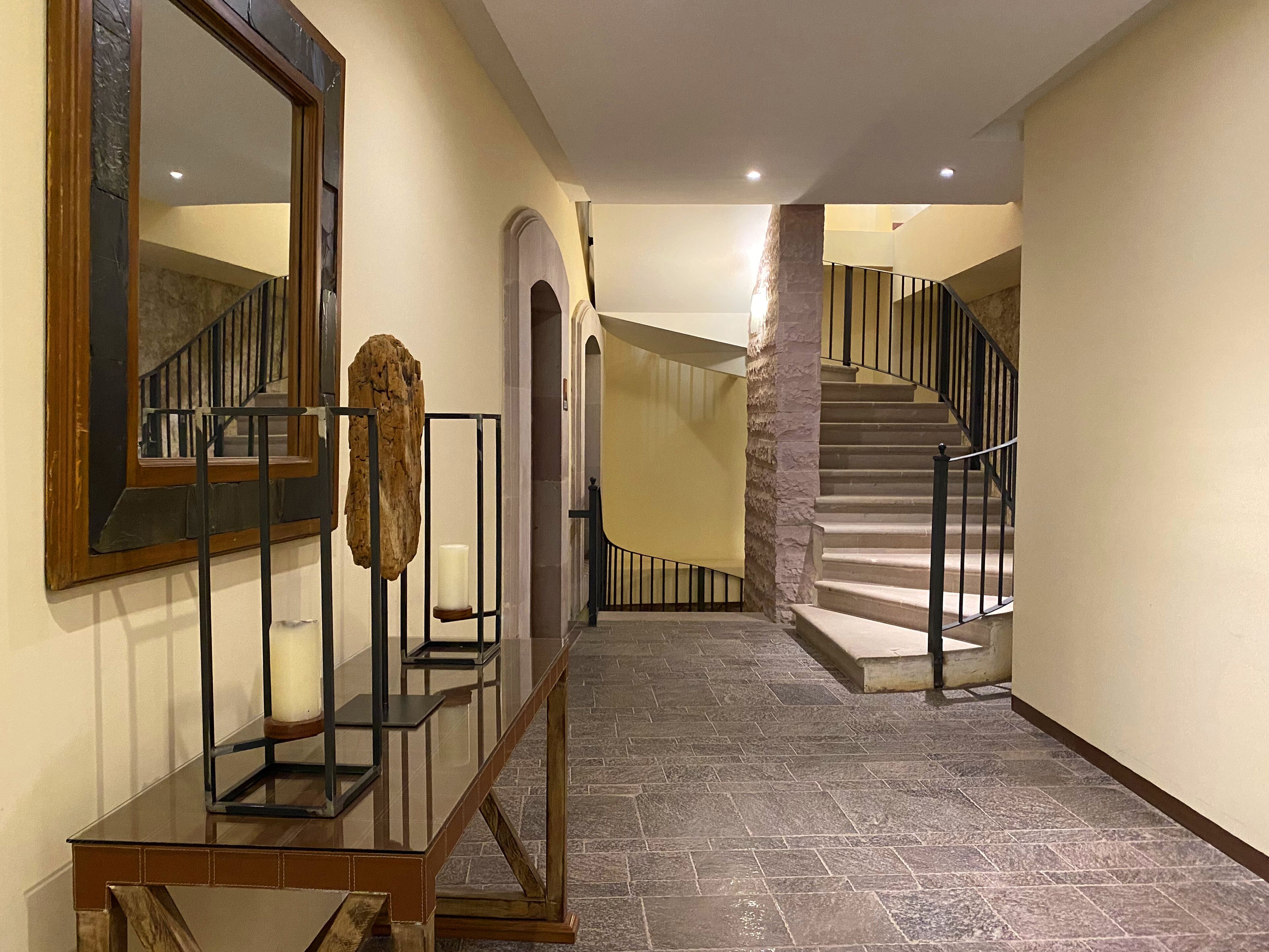 Un pasillo con una escalera, un banco de madera, un espejo y un pilar de piedra.
