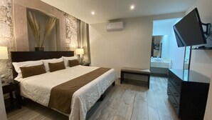 Quarto de hotel com uma cama grande, mesa de cabeceira com abajur, pequeno banco, TV fixada na parede e parede de destaque em pedra.