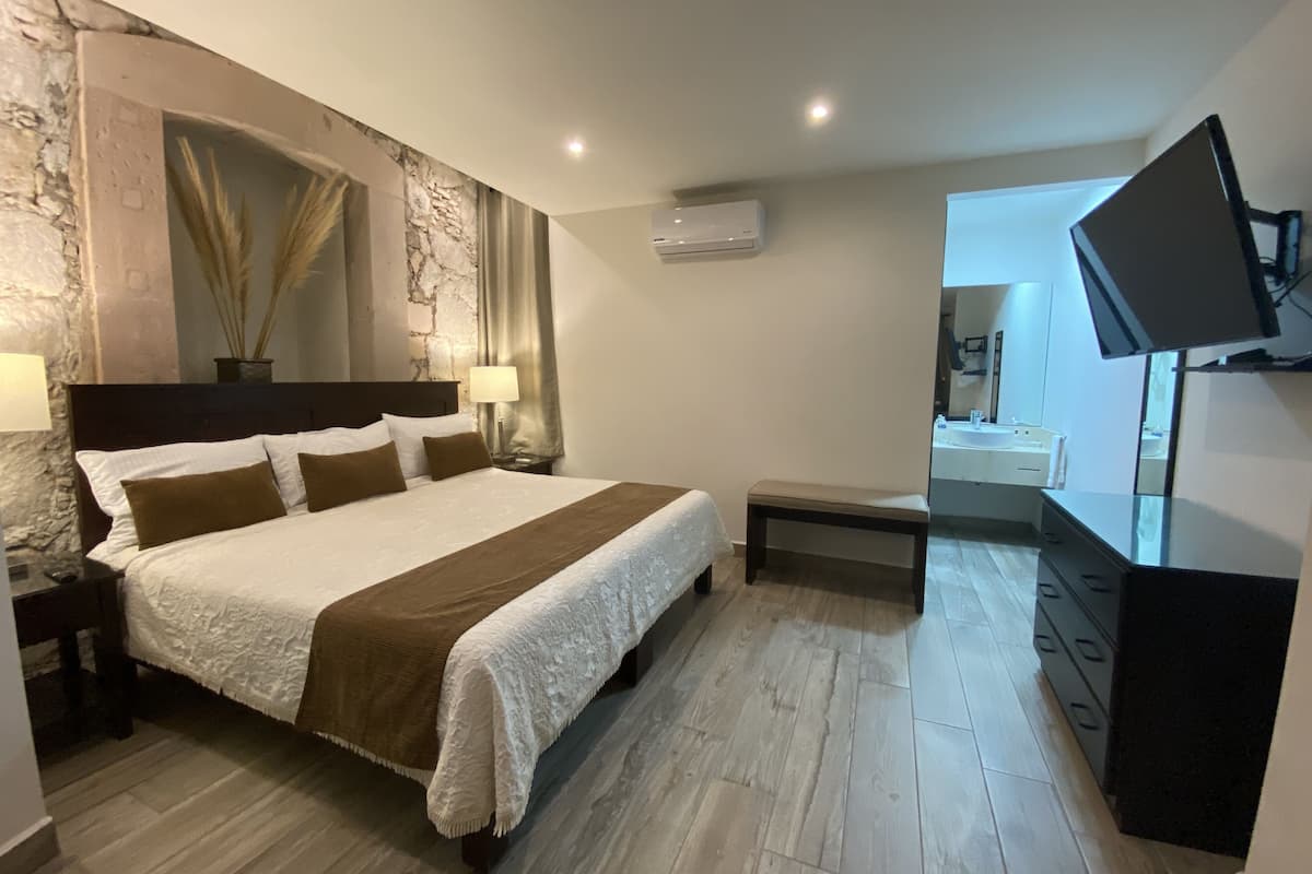 Una habitación de hotel con una cama grande, una mesita de noche con lámpara, un pequeño banco, un televisor montado en la pared y una pared de piedra decorativa.
