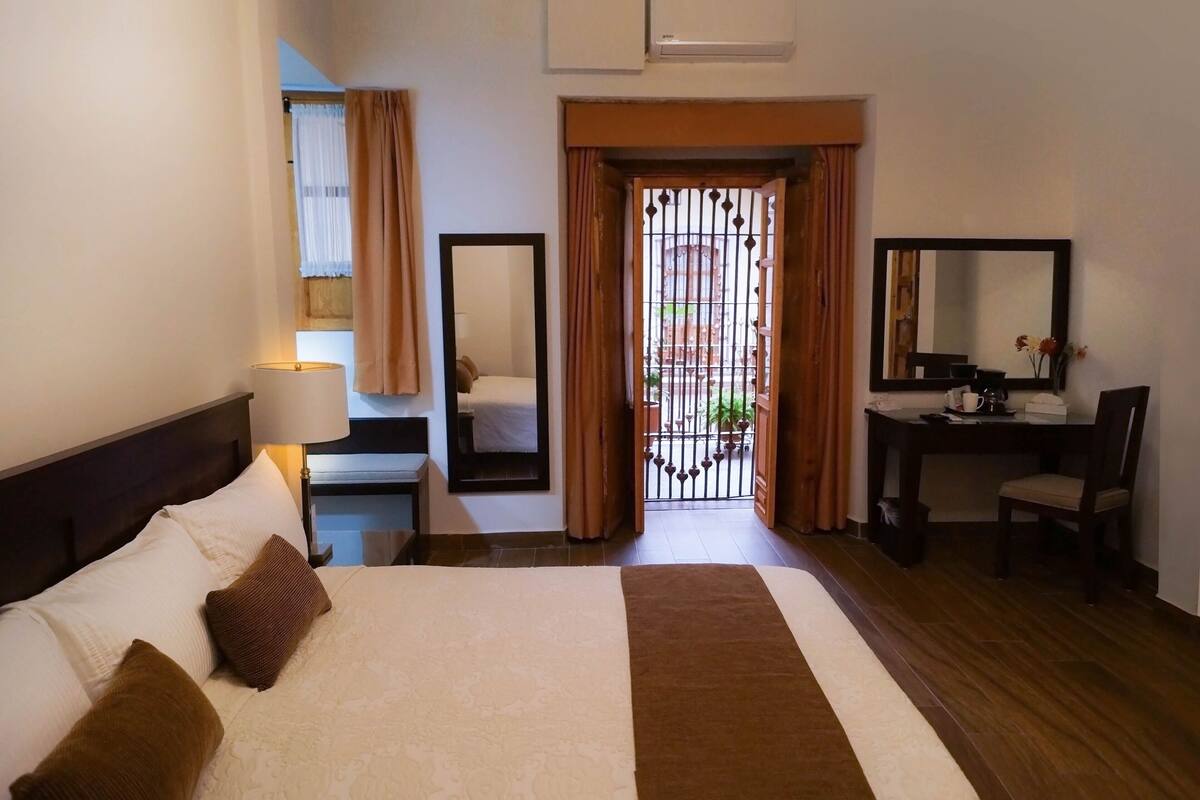 Habitación de hotel con cama, mesita de noche, lámpara, espejo, escritorio, silla y vistas al exterior a través de una reja.