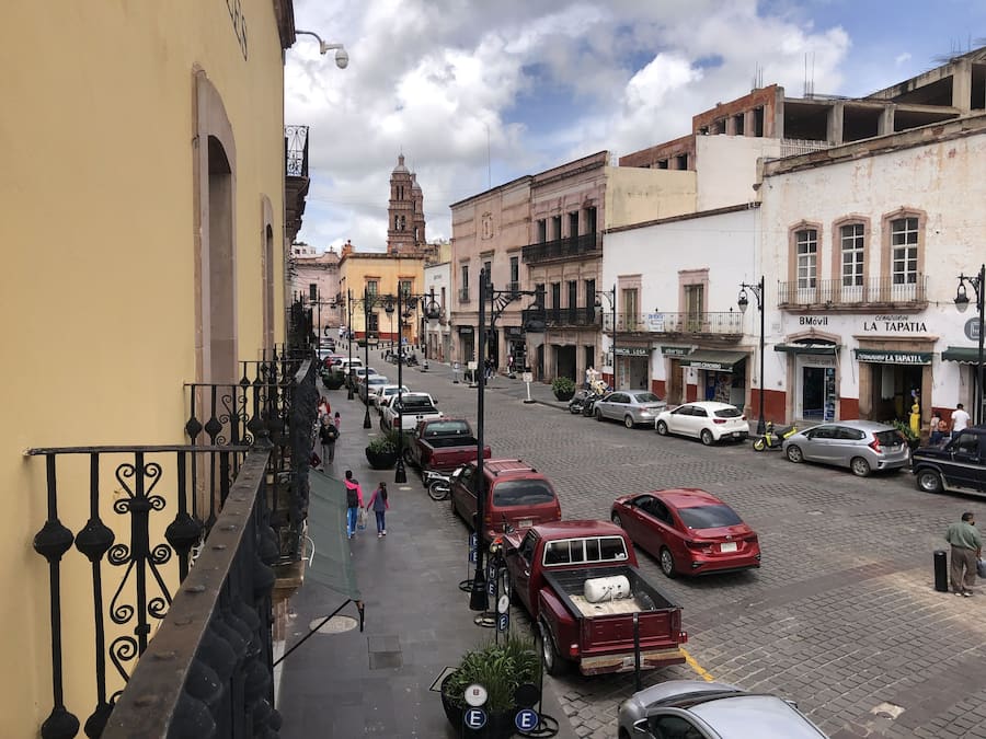 Vista de la calle con edificios, coches aparcados y peatones.
