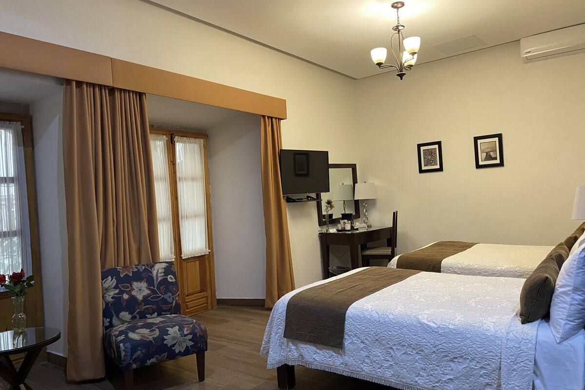 Habitación de hotel con dos camas, un escritorio, una silla, un televisor y un ventanal con cortinas.