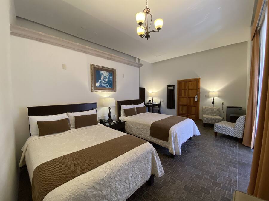 Une chambre d’hôtel avec deux lits, une armoire en bois, un bureau avec une chaise et un lustre.