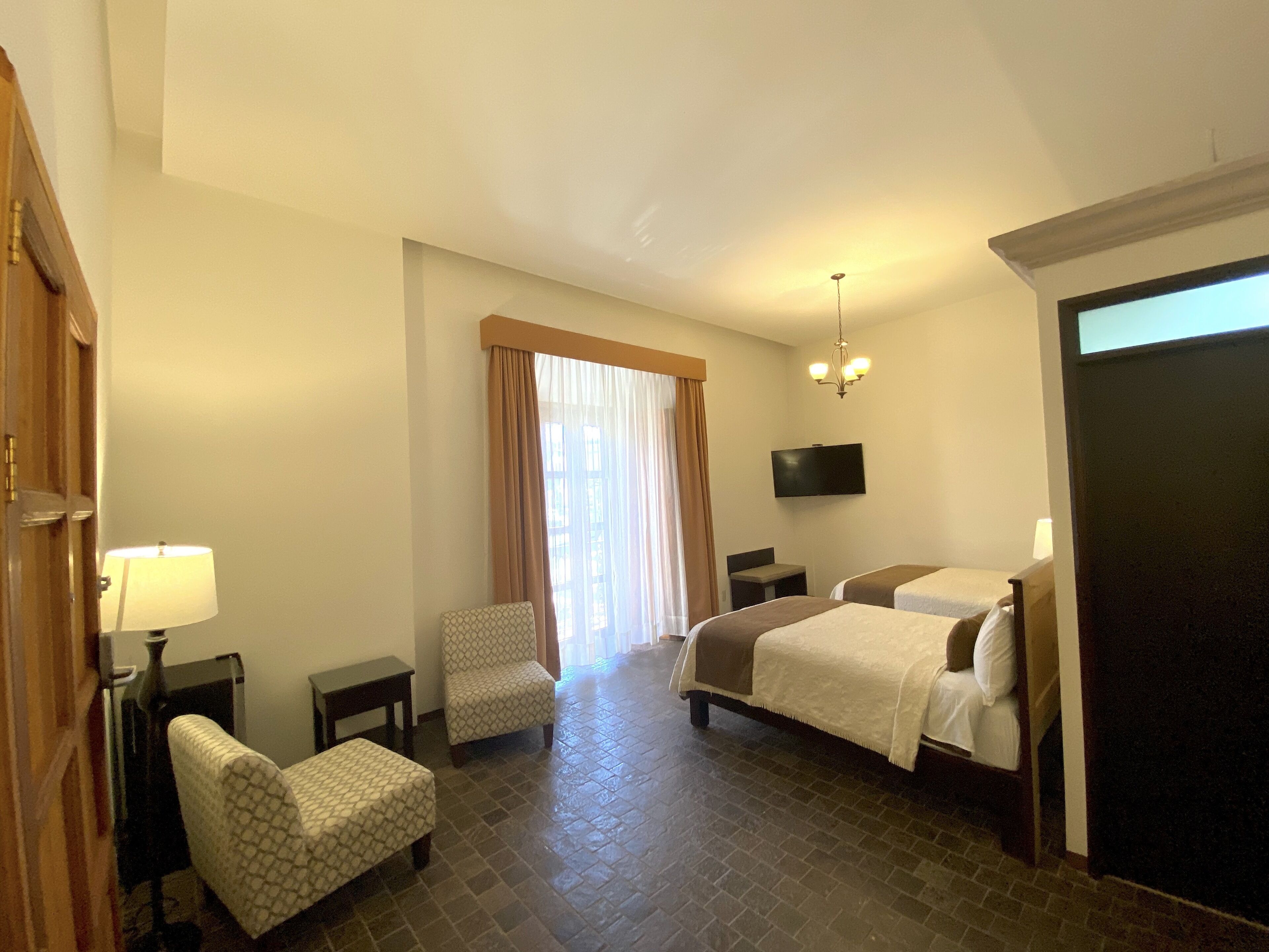 A hotel room with a bed, two chairs, a desk, a TV, and a window with curtains.