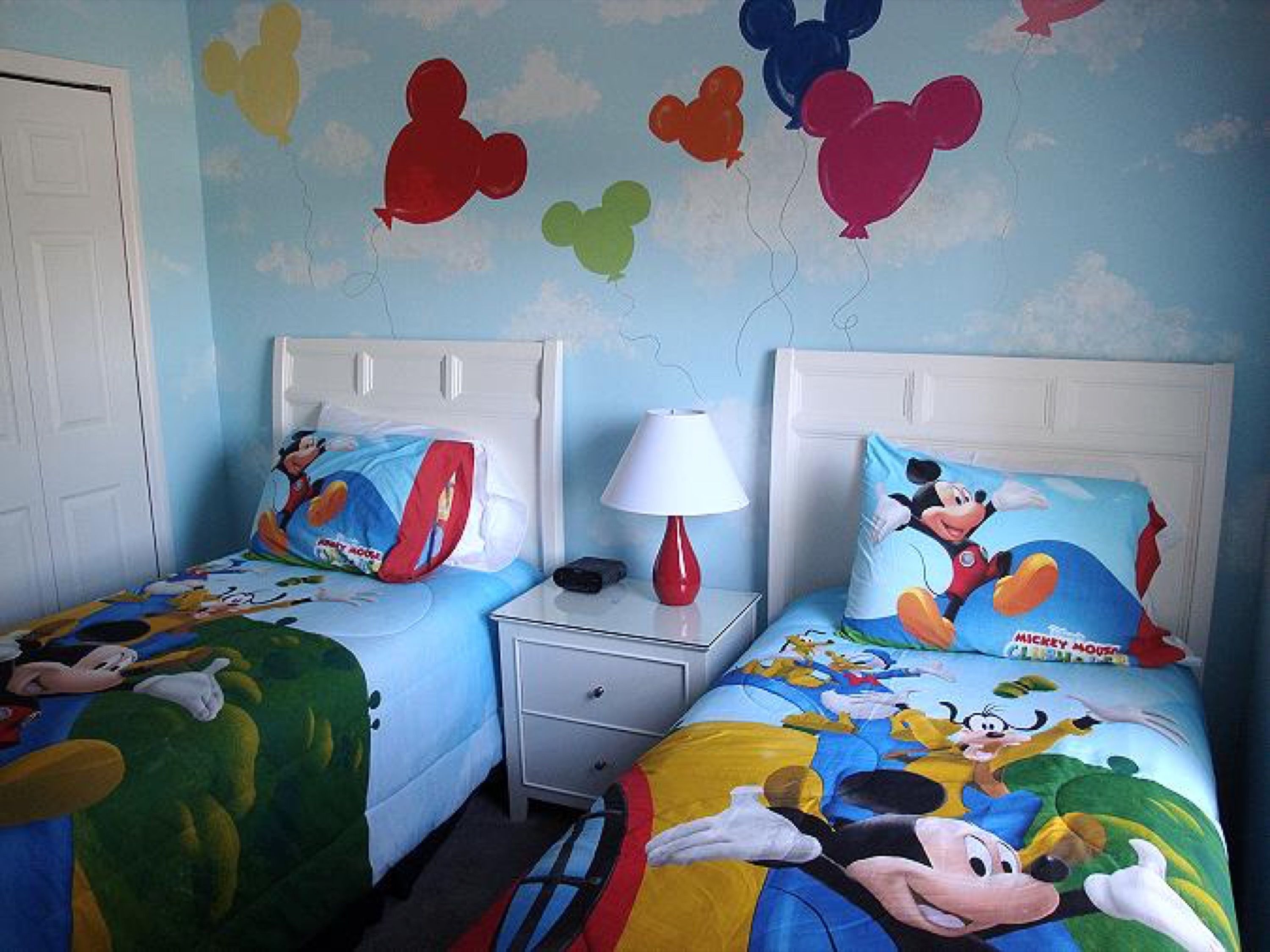 Ein Kinderzimmer mit zwei Betten, beide mit Bettwäsche im Mickey Mouse Design. An einem Nachttisch steht eine Lampe und ein kleines Schubfach.