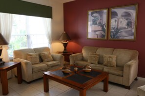 A living room with two beige sofas, a wooden coffee table, and a lamp. There are two framed pictures on the wall. - 7BR 4.5BA Home in Emerald Island Resort by CV-2708 (Kissimmee)
