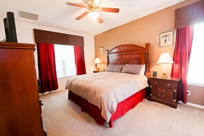 A bedroom with a large bed, a wooden dresser, a nightstand, a ceiling fan, and a window with red curtains.