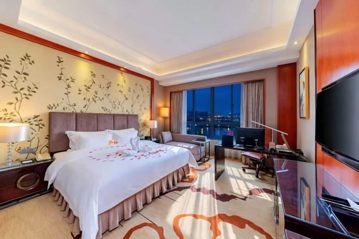 Ein Hotelzimmer mit einem großen Bett, einem Schreibtisch mit Computer, einem Fernseher und Blick auf die beleuchtete Stadt.