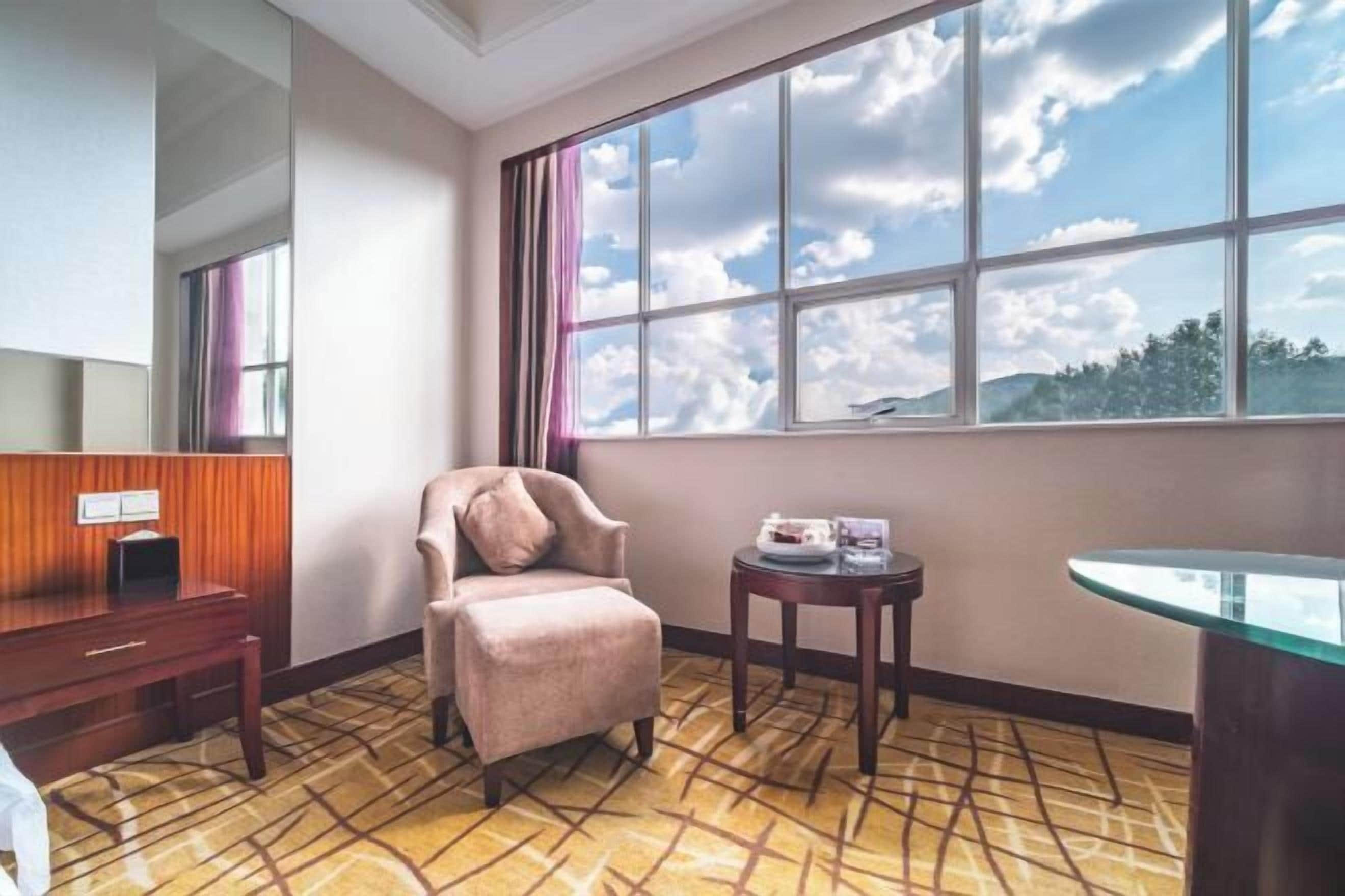 A hotel room with a large window, a chair, a small table, and a desk.