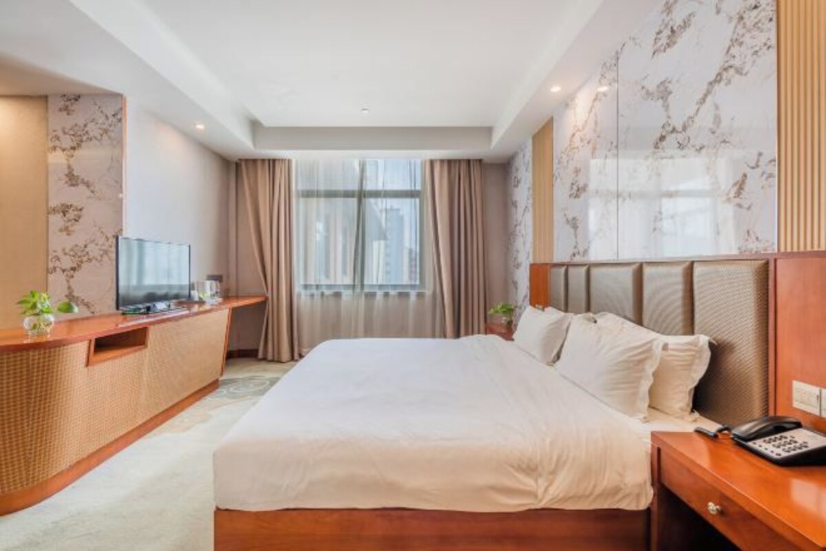 A hotel room with a large bed, a bedside table with a phone, a desk with a TV, and a window with curtains.