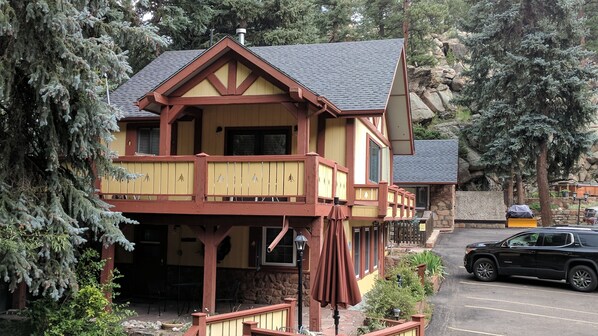 A two-story building with a balcony, stone foundation, and a parked SUV in the driveway.