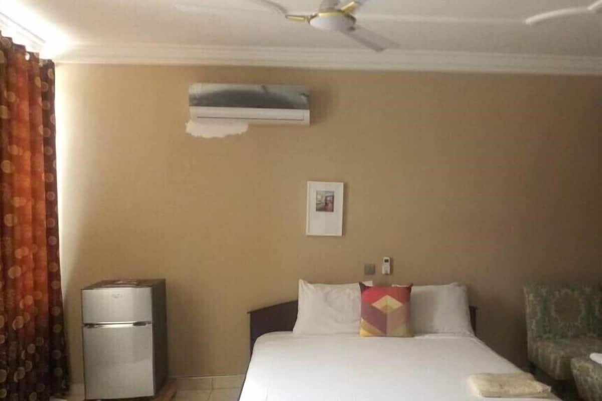 A hotel room with a bed, a small refrigerator, a ceiling fan, an air conditioning unit, a picture on the wall, and a patterned chair.