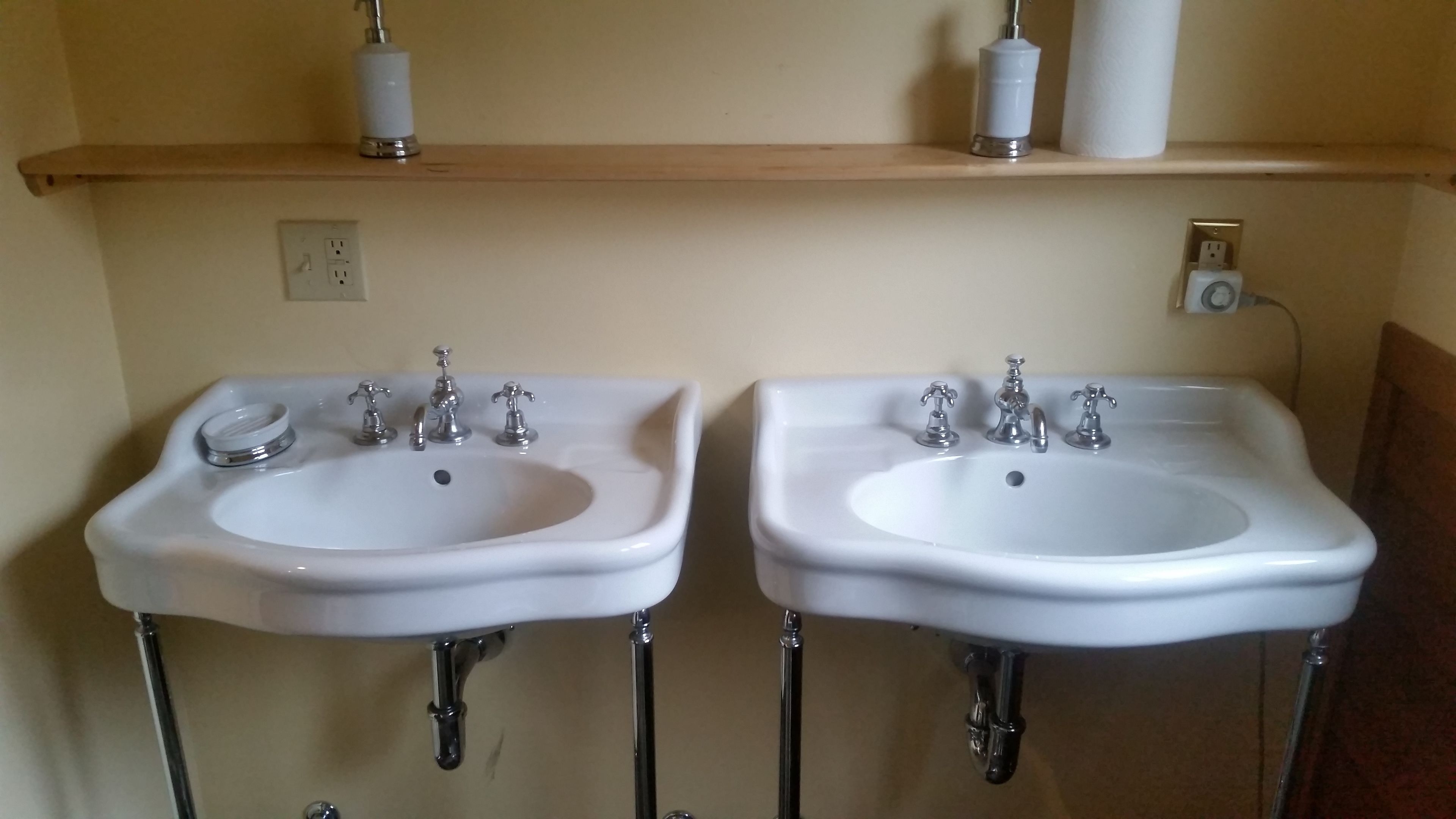 Two white bathroom sinks with chrome faucets, a wooden shelf above them holding toilet paper rolls, and a wall socket.