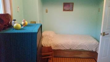 Una habitación pequeña con una cama, una mesa con mantel azul y una silla. Hay un cuadro en la pared y una puerta a la derecha.