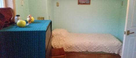 A small room with a bed, a table with a blue tablecloth, and a chair. There is a picture on the wall and a door on the right.