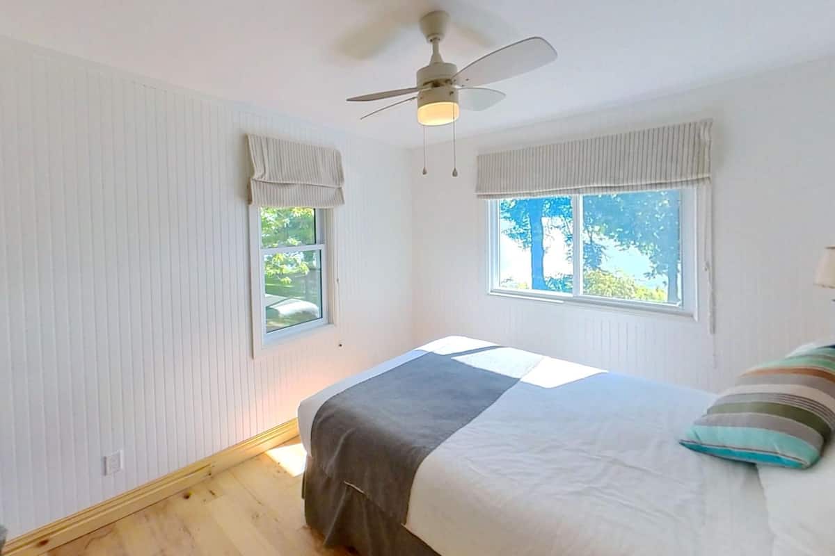 Un dormitorio con una cama, un ventilador de techo, dos ventanas con cortinas y una silla.