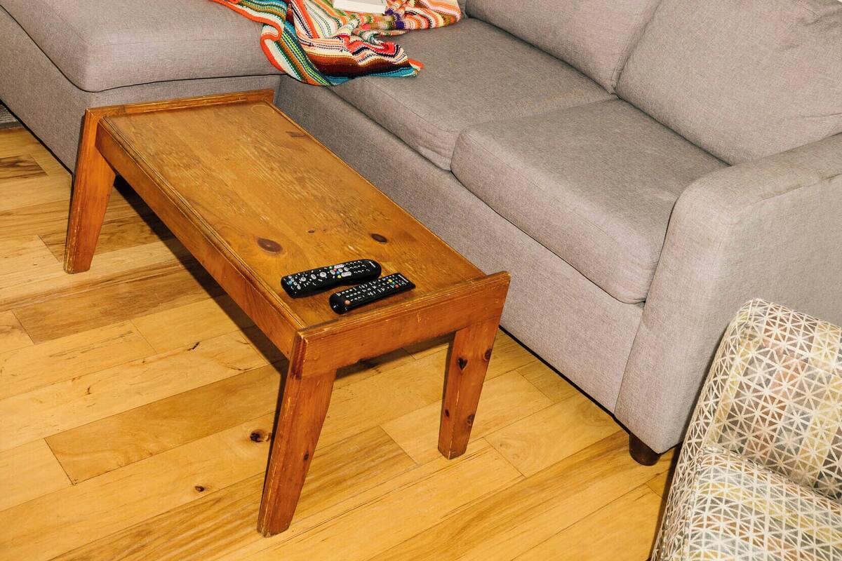 Una sala de estar con un sofá gris, una mesa de centro de madera y una manta colorida. Hay un cuadro en la pared y una ventana con vistas a zonas verdes.