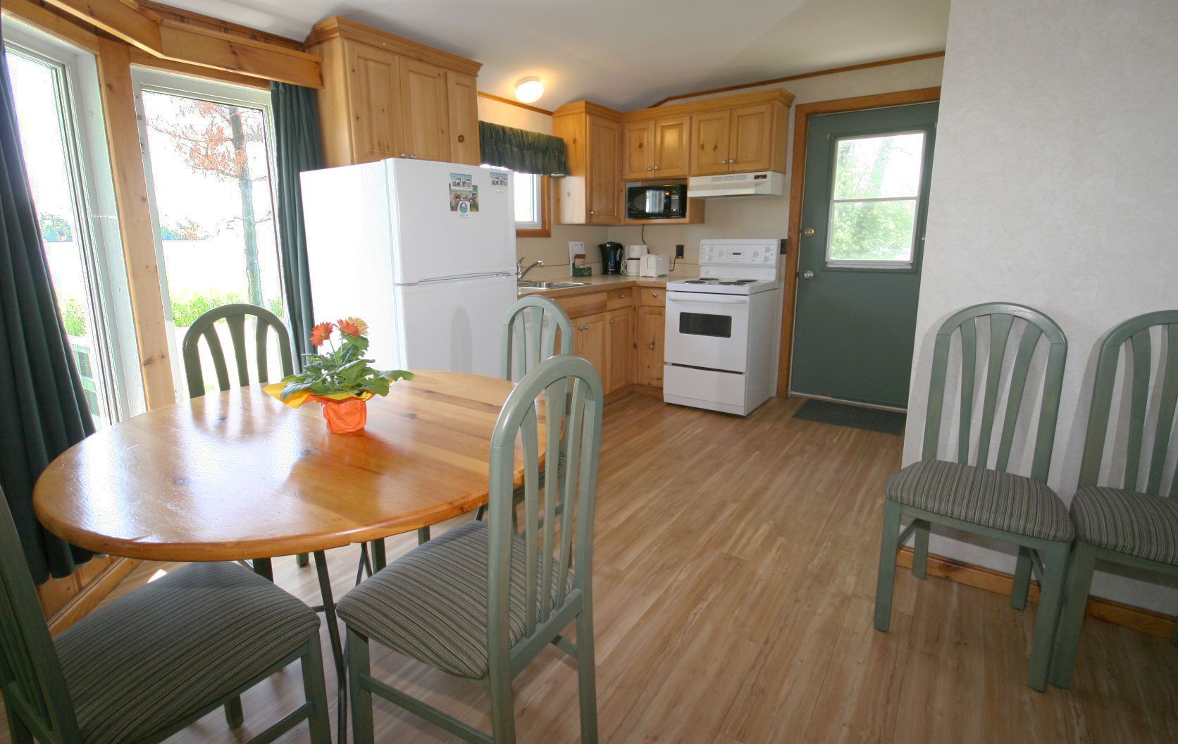 A compact kitchen with wooden cabinets, a refrigerator, a microwave, and a stove. There is a dining table with chairs and a potted plant on it.