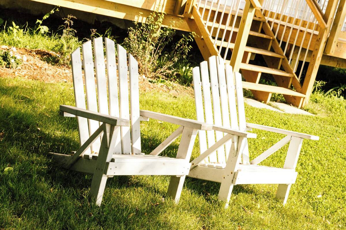 Dos sillas Adirondack sobre un césped frente a una casa con terraza de madera y una escalera que lleva a un ventanal del segundo piso.