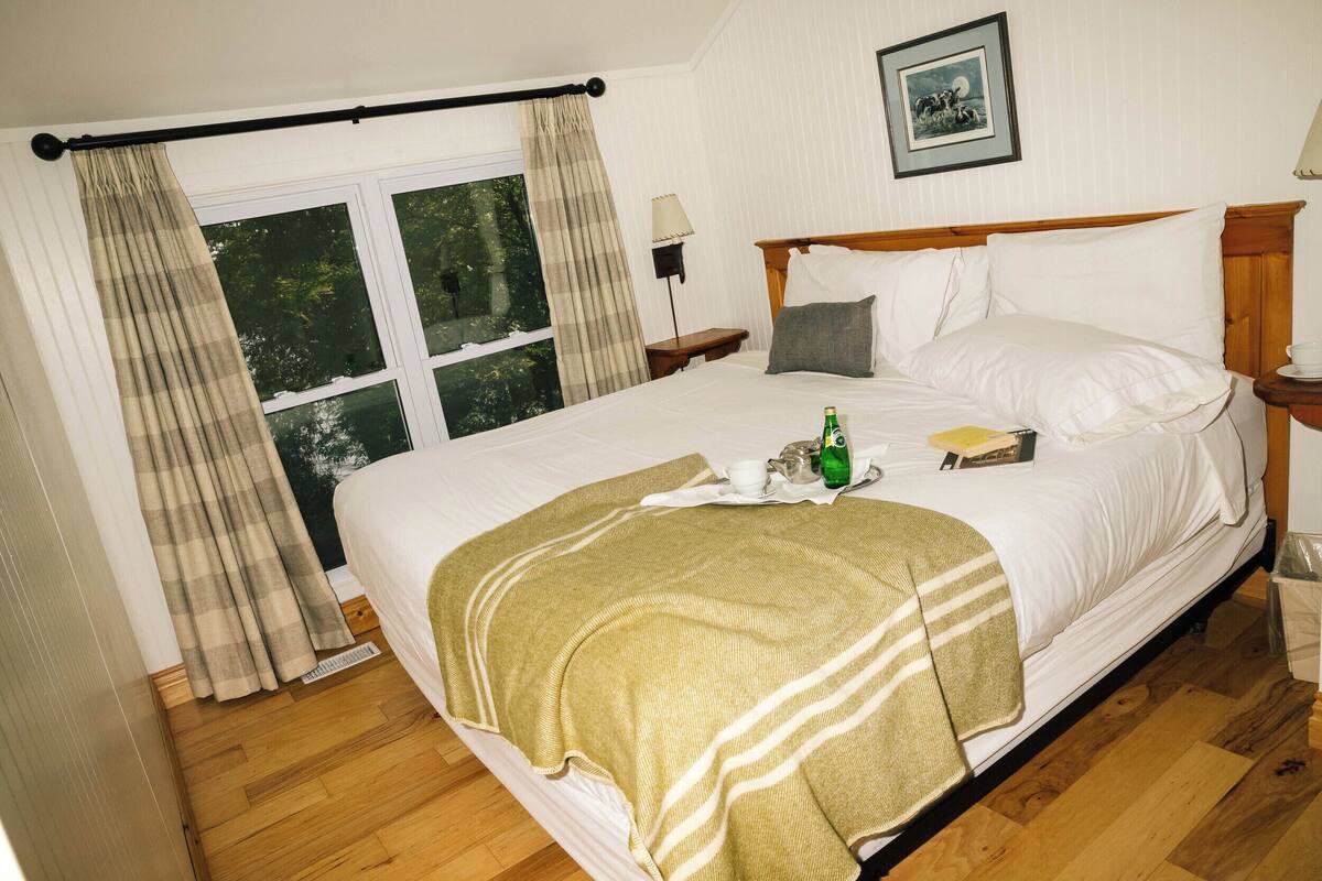 Una cama bien hecha con un edredón a rayas, una mesita de noche con una lámpara y una ventana con cortinas.