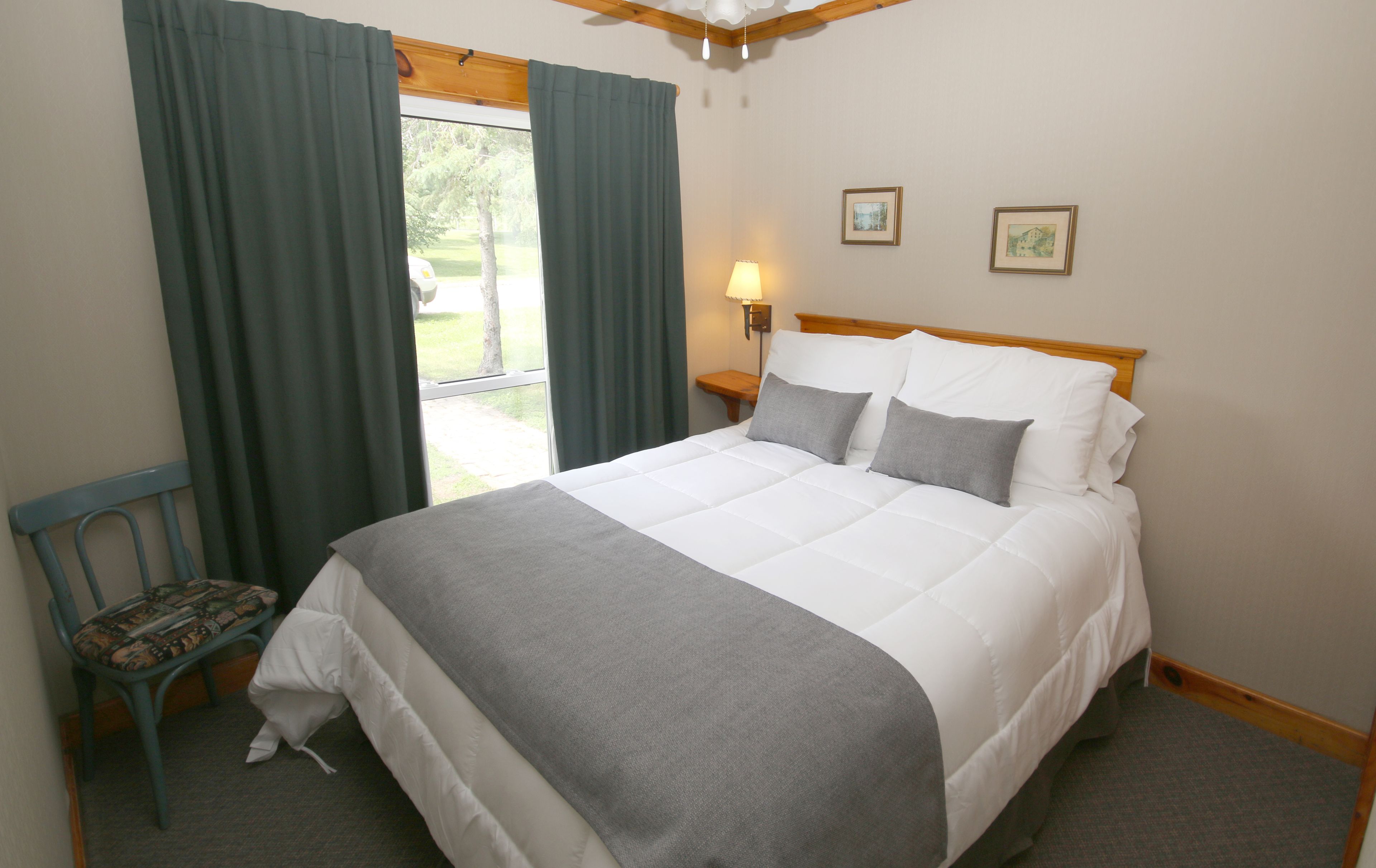 A neatly made bed with a grey blanket and pillows, a wooden headboard, a nightstand with a lamp, a chair with a patterned cushion, and a window with curtains.